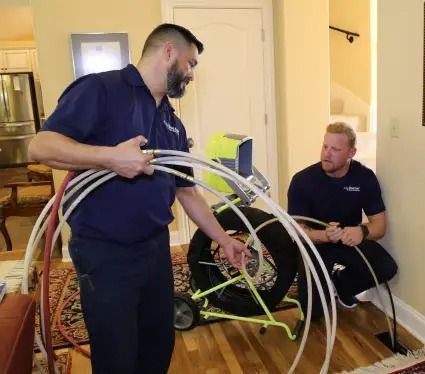 Two men are working on a pipe in a living room.