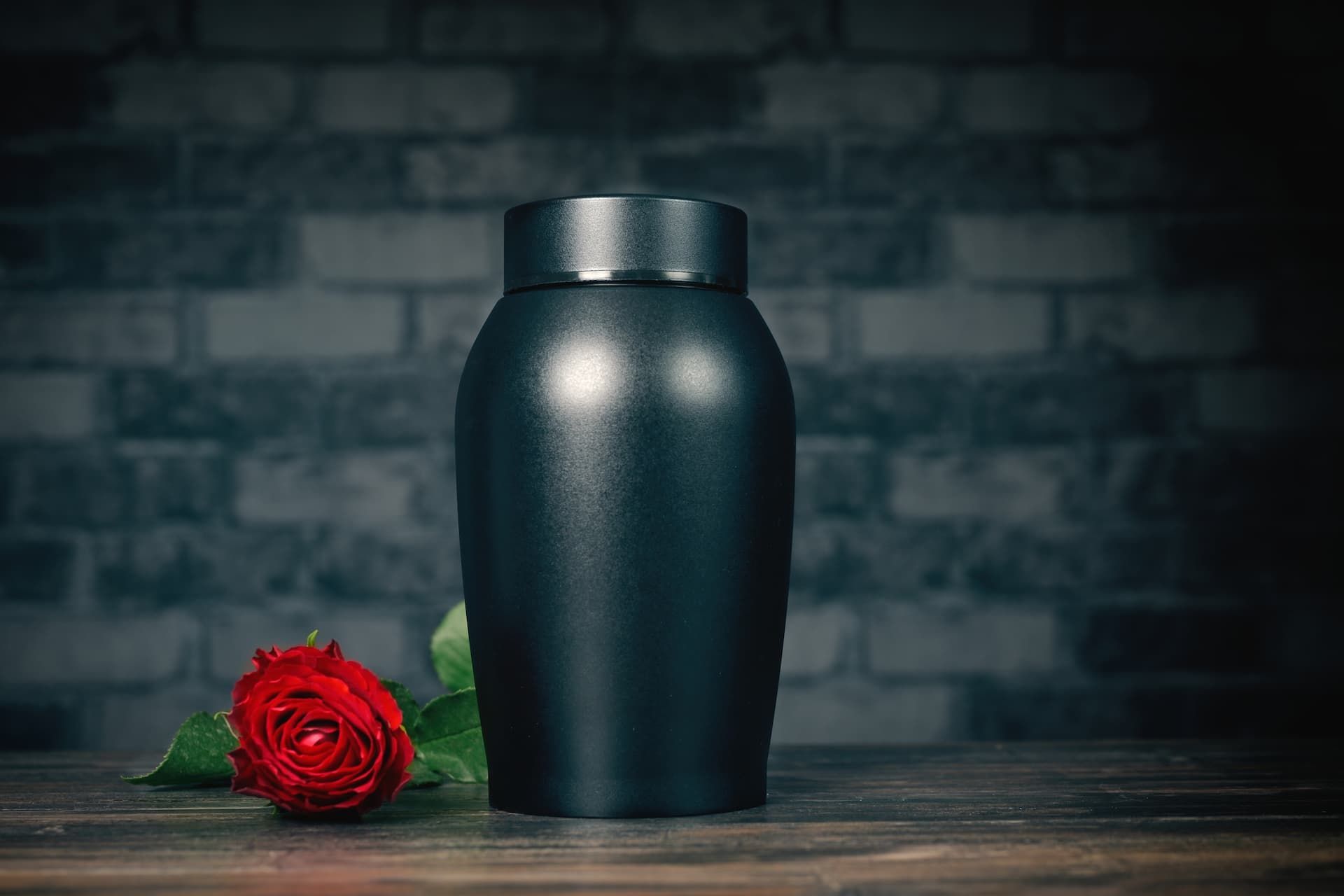 Black urn and red rose on a wooden surface in front of a brick wall.