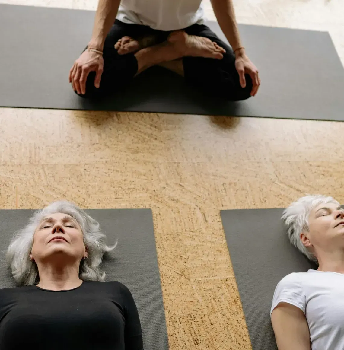 Three people lying on yoga mats in a studio, eyes closed, in a relaxation pose