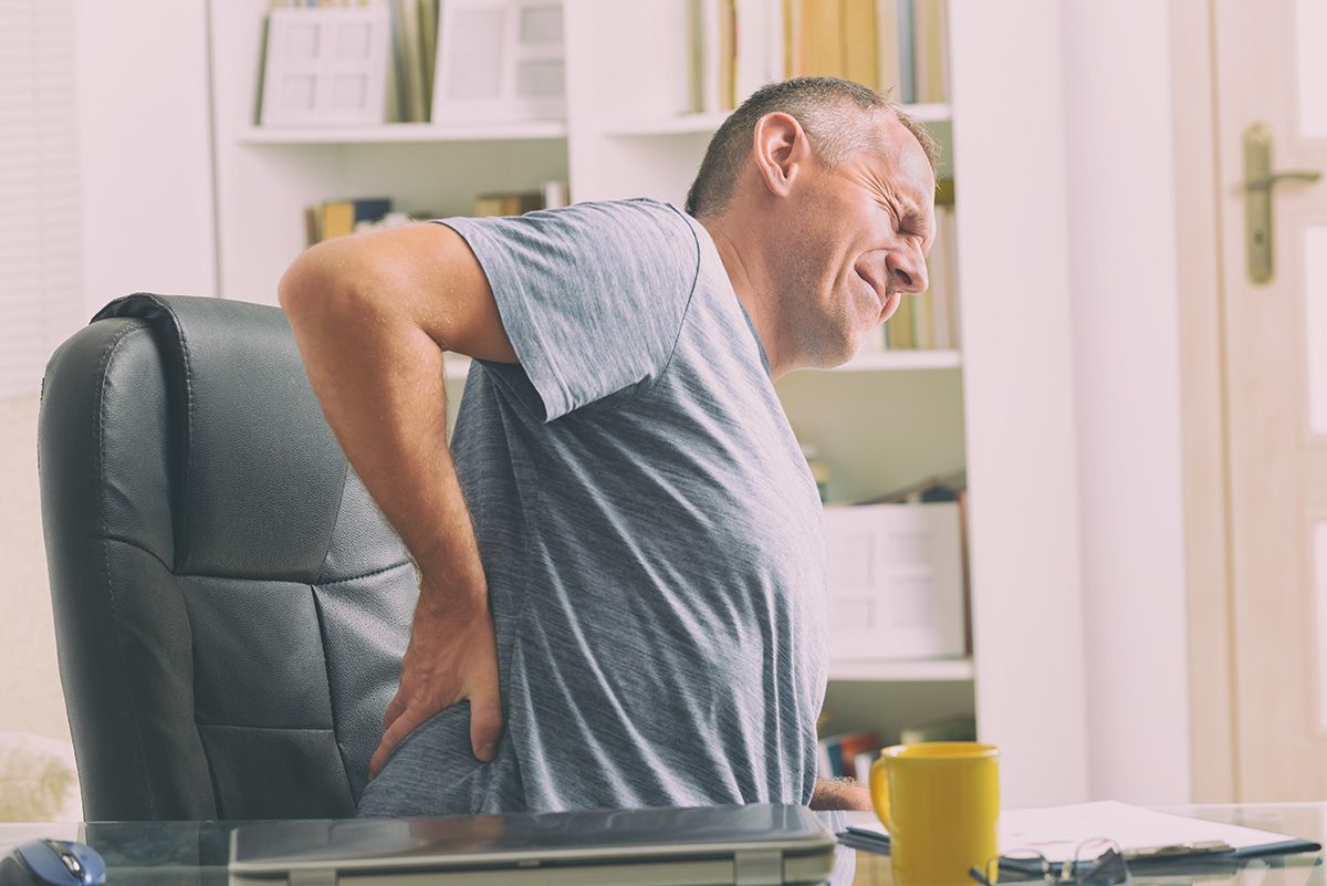 Man holding his lower back in pain while seated at a desk in a bright office