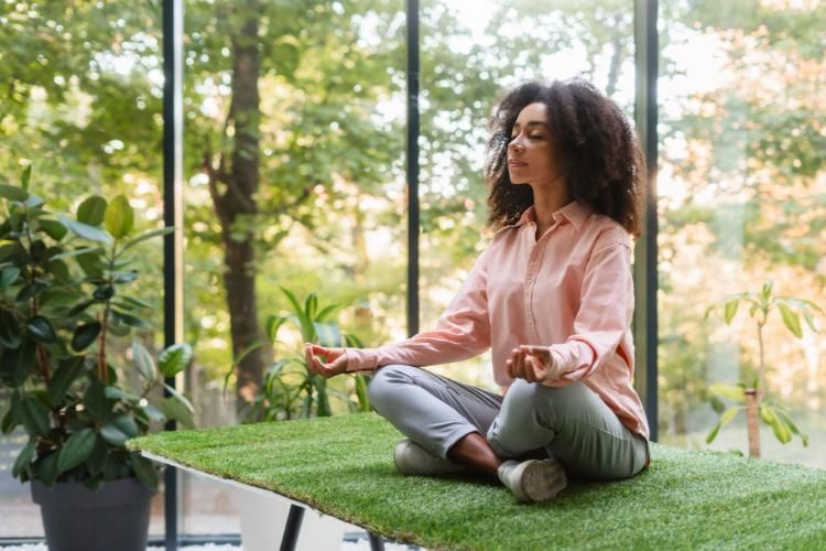 Person meditating cross-legged on a grassy rooftop beside plants and large windows.
