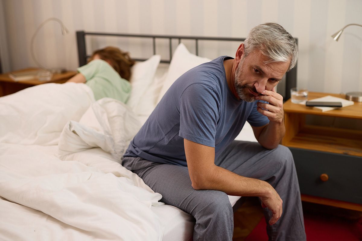 Man sitting on the edge of a bed, looking pensive, with a woman resting in the background and a bedside table nearby