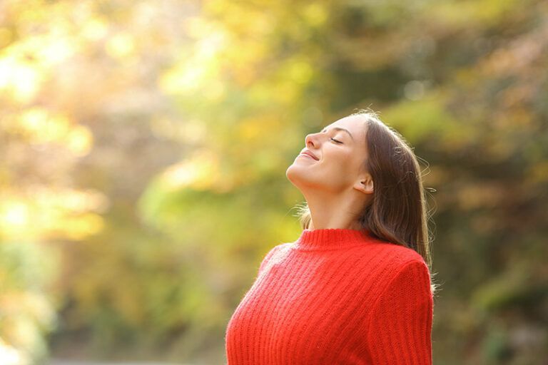 Woman in a red sweater smiling with eyes closed in a sunlit autumn park