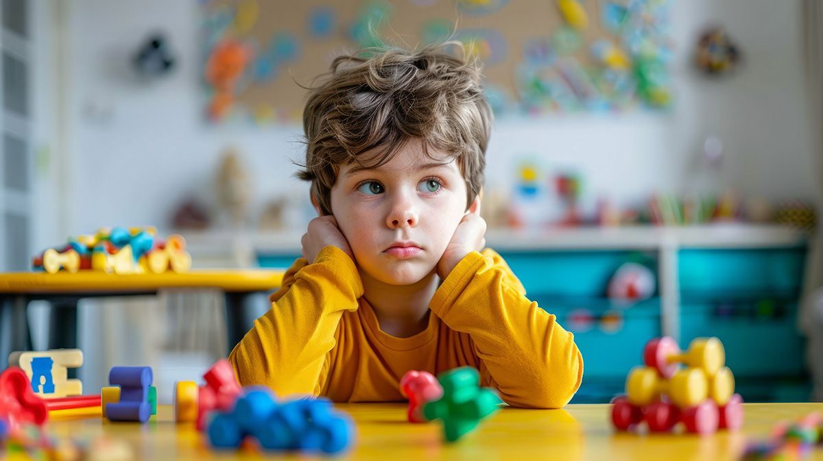 Child with curly hair resting chin on hands at a colorful toy table in a playroom