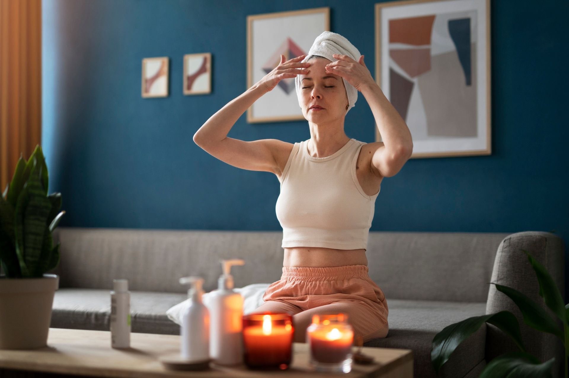 Person sitting on a couch applying a towel to their head, with candles and bottles on a coffee table in a blue living room.
