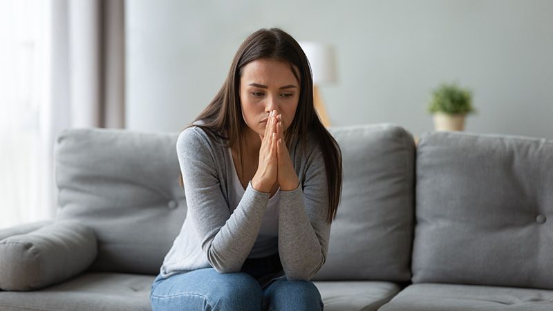 Woman sitting on a gray couch with hands clasped, looking down thoughtfully in a living room