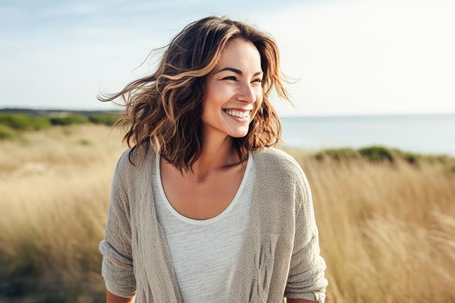 Smiling woman in a beige cardigan standing in tall grass by the coast