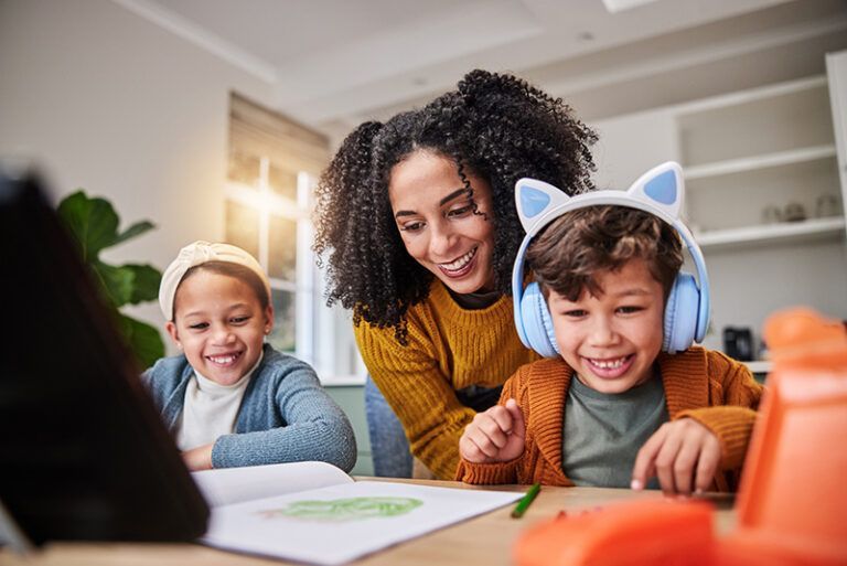 Woman helping two children with homework at a table, smiling indoors with books and pencils.