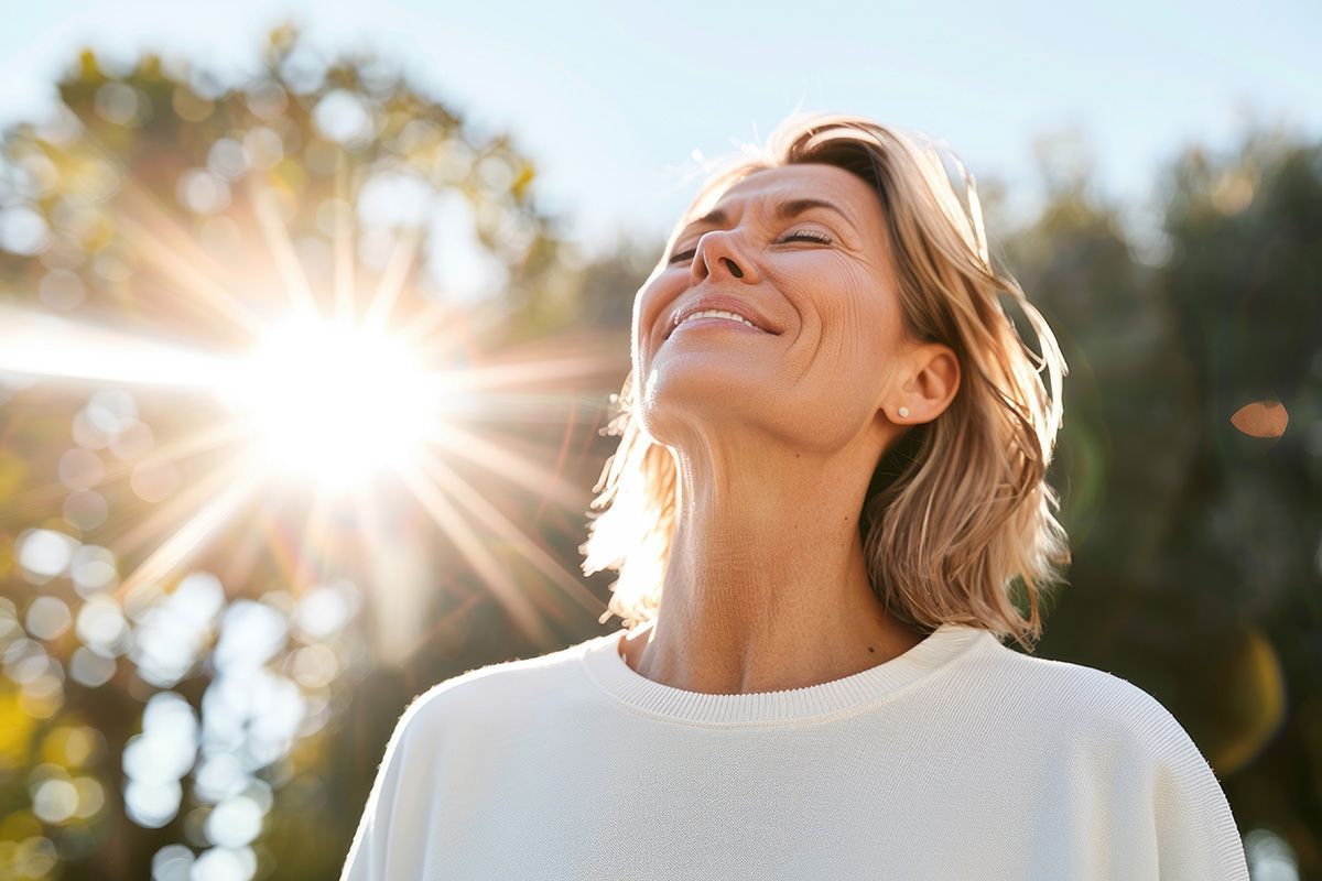 Smiling woman in a white sweater enjoying sunlight outdoors with trees in the background