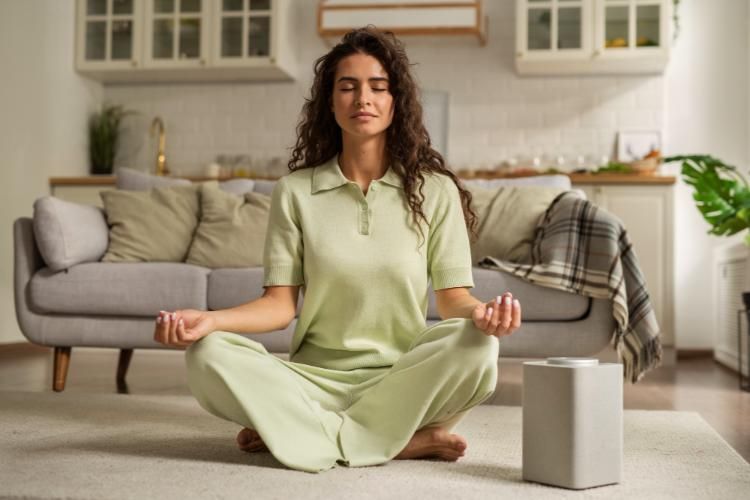 Person meditating cross-legged in a bright living room with a sofa and plaid blanket nearby
