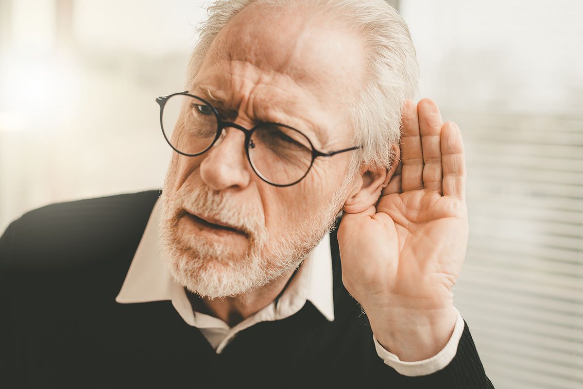 Older man with glasses cupping his ear, listening intently against a light background