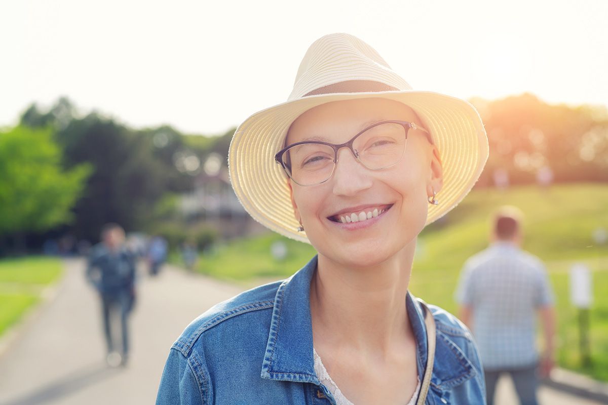 Smiling woman in a straw hat and denim jacket outdoors in warm sunlight, with people blurred in the background