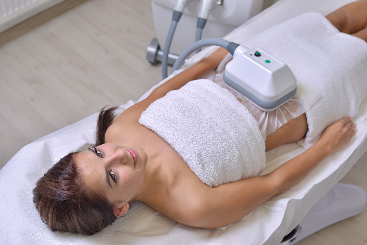 Woman lying on a spa treatment bed with a device on her abdomen and a towel wrapped around her torso