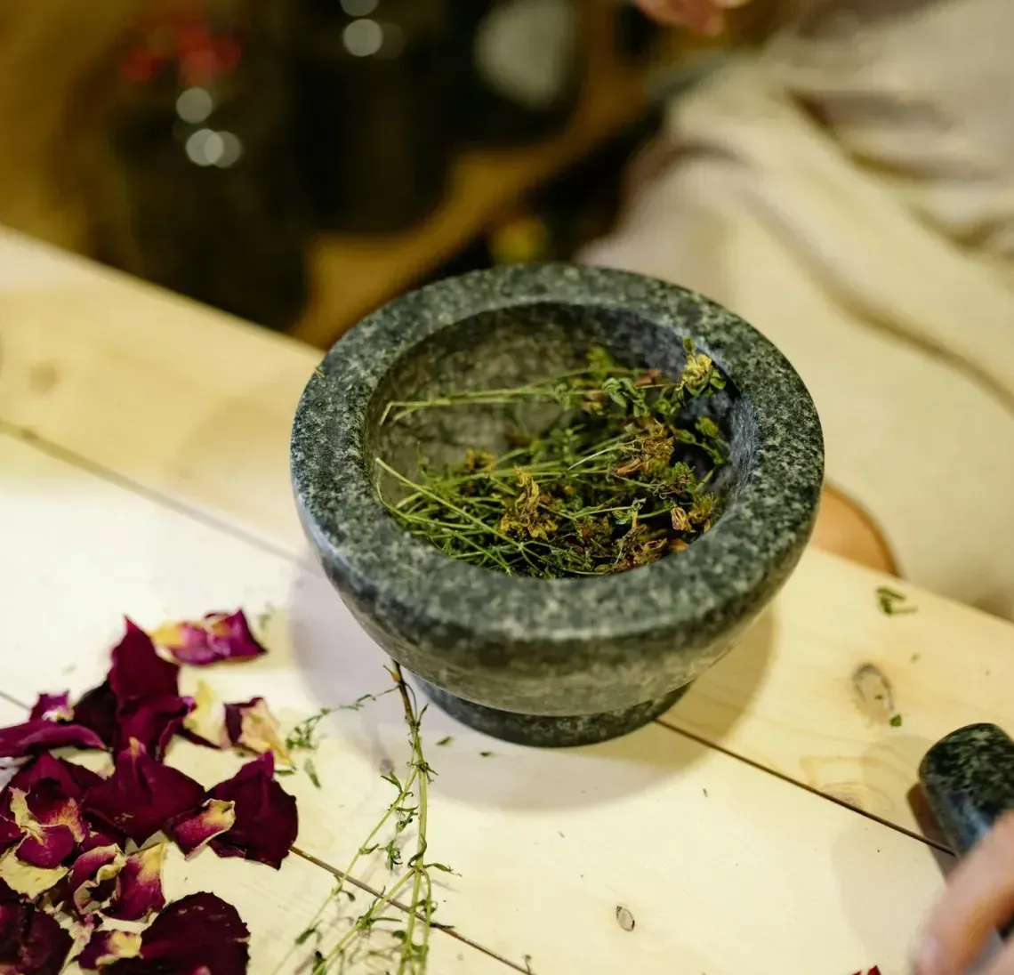 Mortar and pestle with crushed herbs on a table, with scattered rose petals and a hand nearby