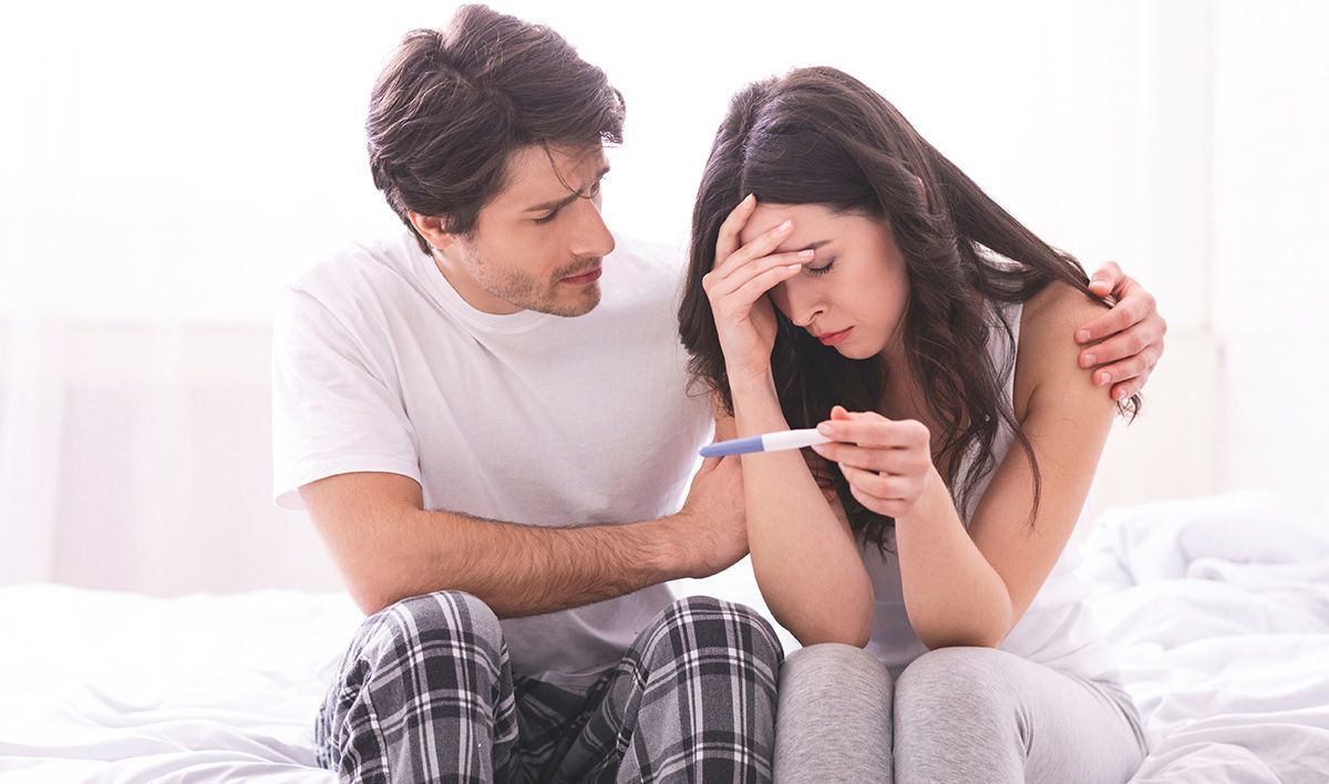 A couple sits on a bed, looking upset at a pregnancy test, with the man comforting the woman.