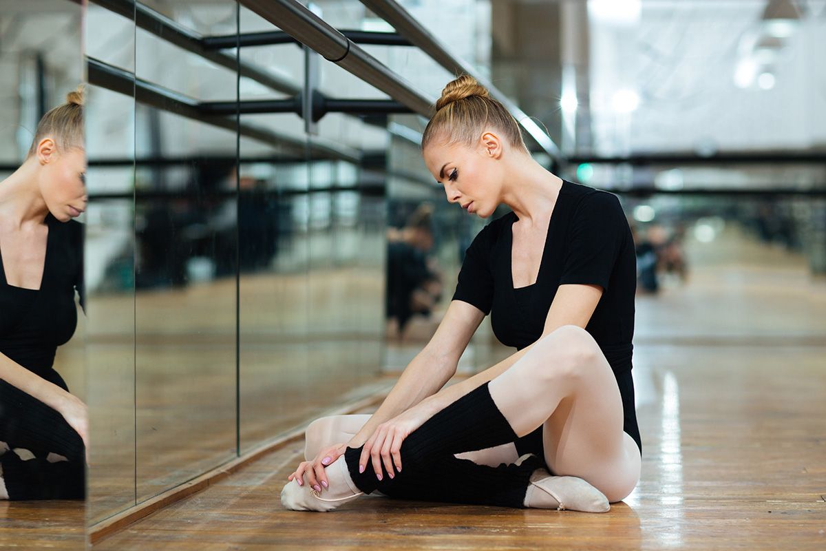 Dancer stretching on wooden studio floor beside mirrored wall, wearing a black outfit and pink ballet shoes