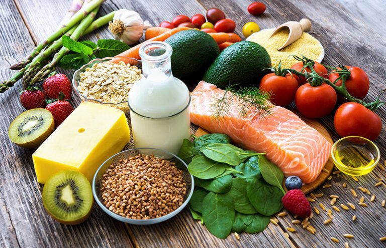 Assorted fresh foods on a wooden table: salmon, vegetables, kiwi, grains, eggs, milk, and oil.
