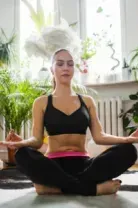 Person meditating cross-legged on a yoga mat in a bright plant-filled room
