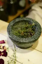 Mortar and pestle with crushed herbs on a table, with scattered rose petals and a hand nearby