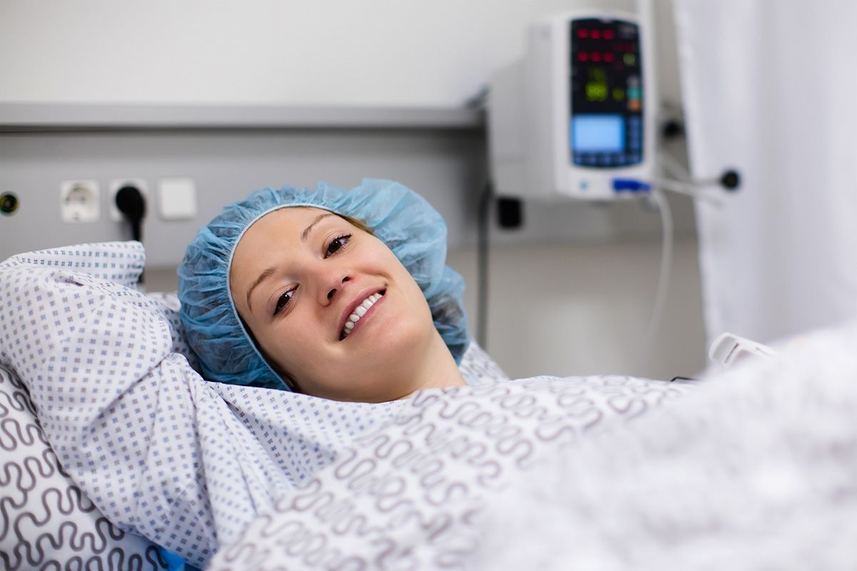 Smiling patient in a hospital bed wearing a blue hair cap, with medical monitor in the background
