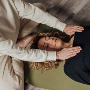 Person receiving a head massage while lying on the floor on a green mat, with another person kneeling nearby