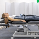 Person lying on a padded exam table in a clinic, with a white privacy screen behind and medical equipment below.