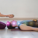 Two people practicing yoga; one kneels holding a singing bowl over the other lying on a mat with crystal bowls nearby