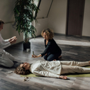 Two people kneel beside a woman lying on the floor in a bright room, as one appears to check her breathing.
