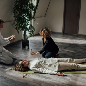 Two people kneel beside a woman lying on the floor in a bright room, as one appears to check her breathing.