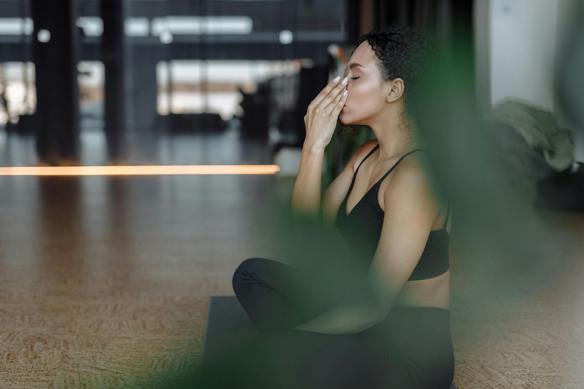 Woman in black workout clothes sitting on a mat, wiping her face in a gym or studio.