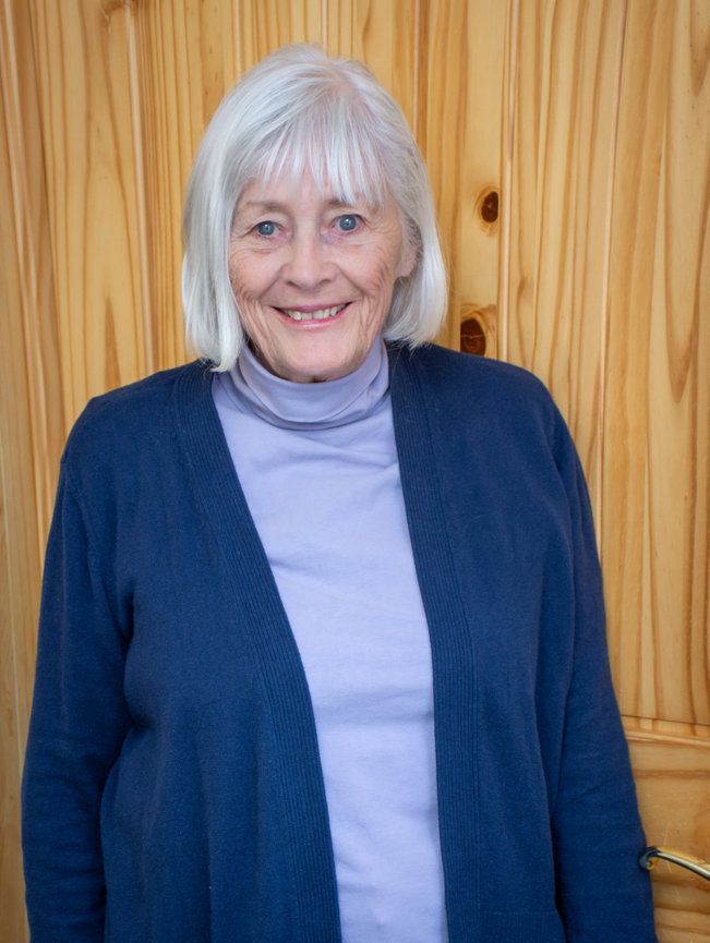 An elderly woman wearing a blue sweater and a purple shirt is smiling in front of a wooden wall.