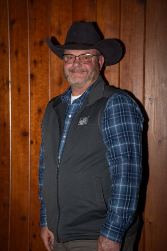 A man wearing a cowboy hat and vest is standing in front of a wooden wall