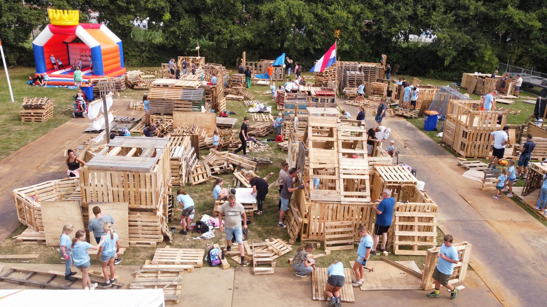 Mensen bouwen constructies van houten pallets op een grasveld, met een springkussen op de achtergrond.