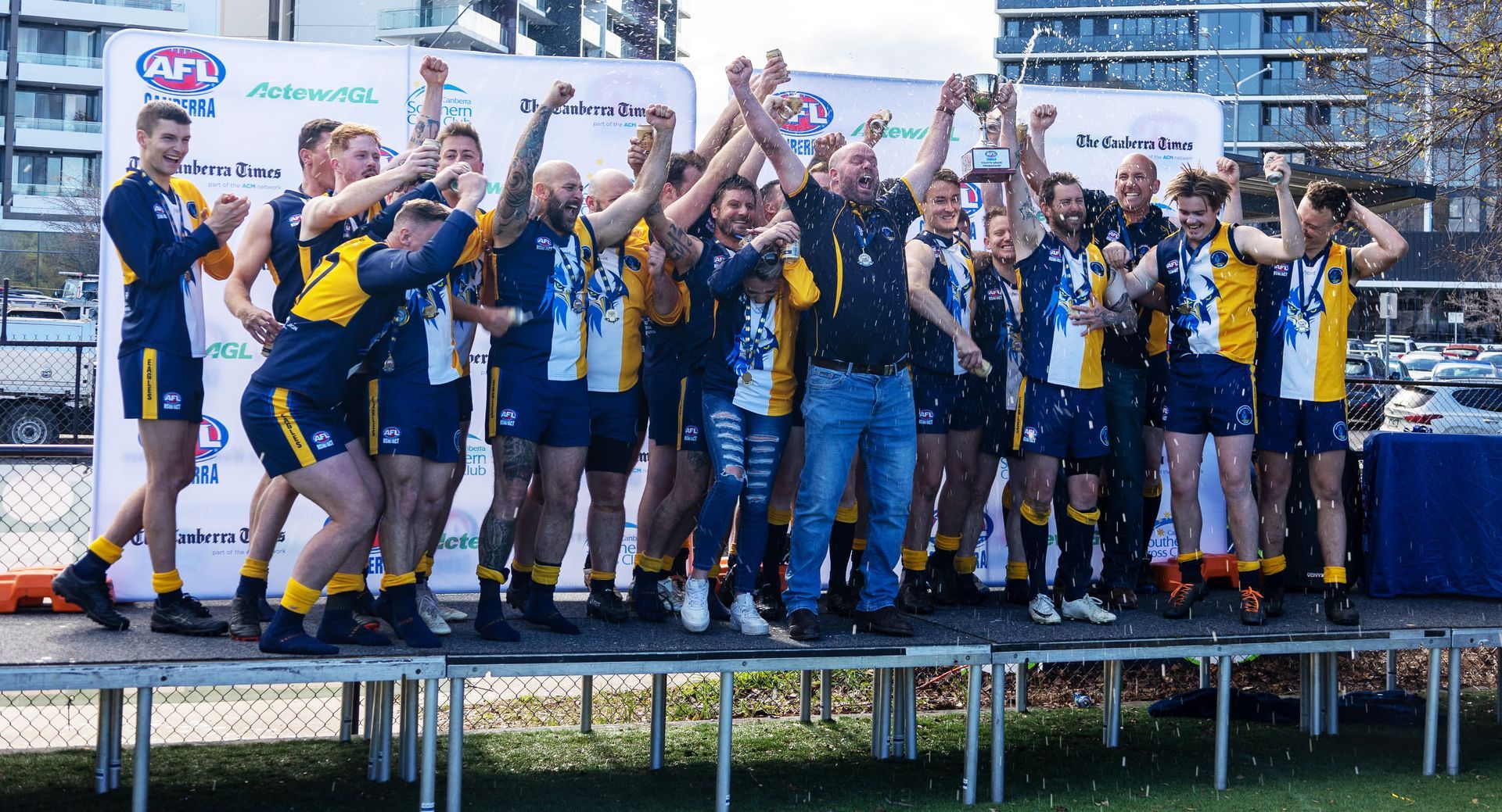 A sports team in blue and yellow uniforms celebrates on a stage, cheering and holding a trophy outdoors.