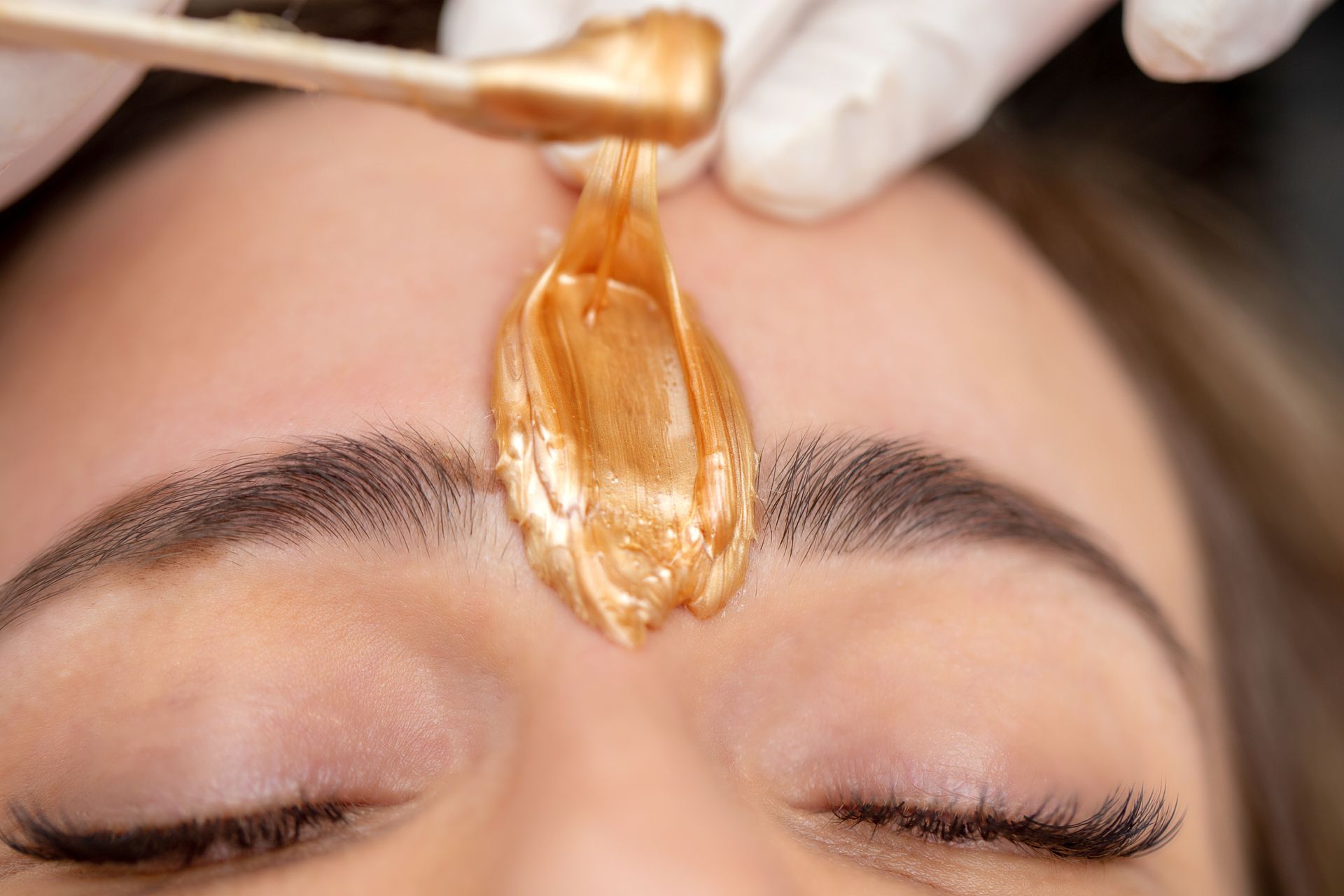 A person's forehead being waxed between their eyebrows. A gloved hand applies golden wax.