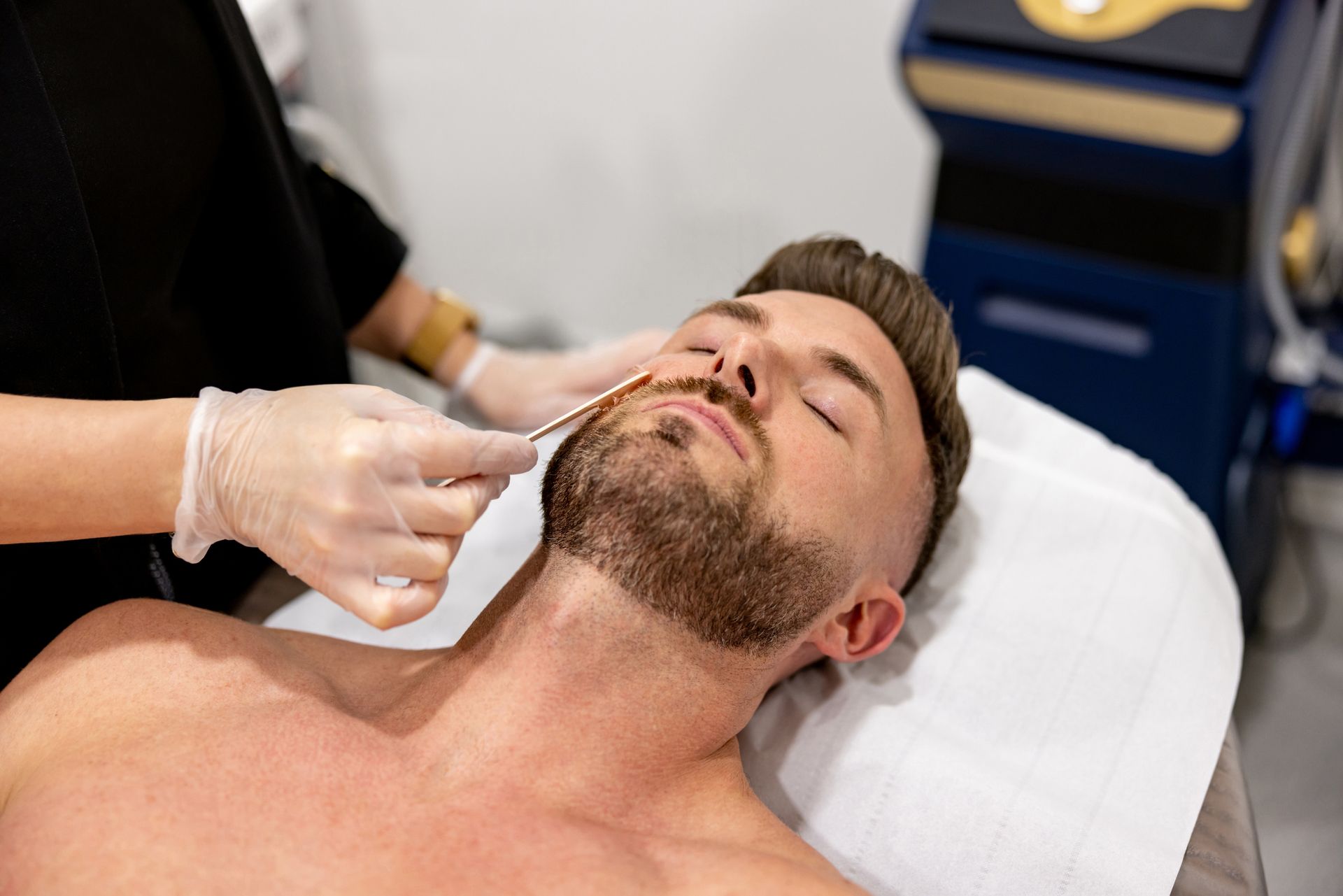 Man receiving facial hair treatment, lying on a white table. A gloved hand applies something to his upper lip in a clinic.