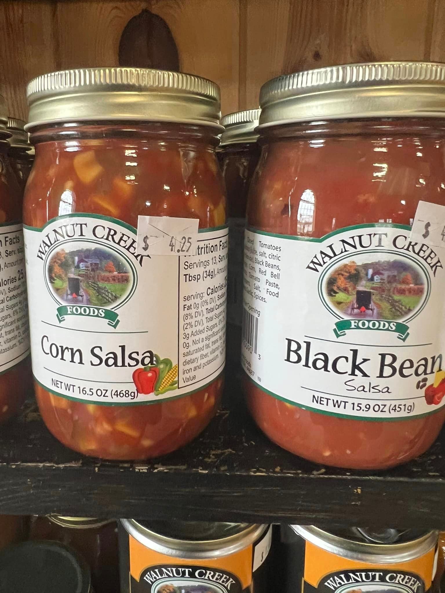Two jars of corn salsa and black bean salsa are sitting on a shelf.