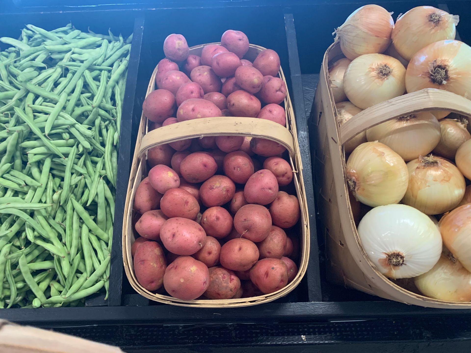 Three baskets of potatoes , green beans and onions are on a shelf.