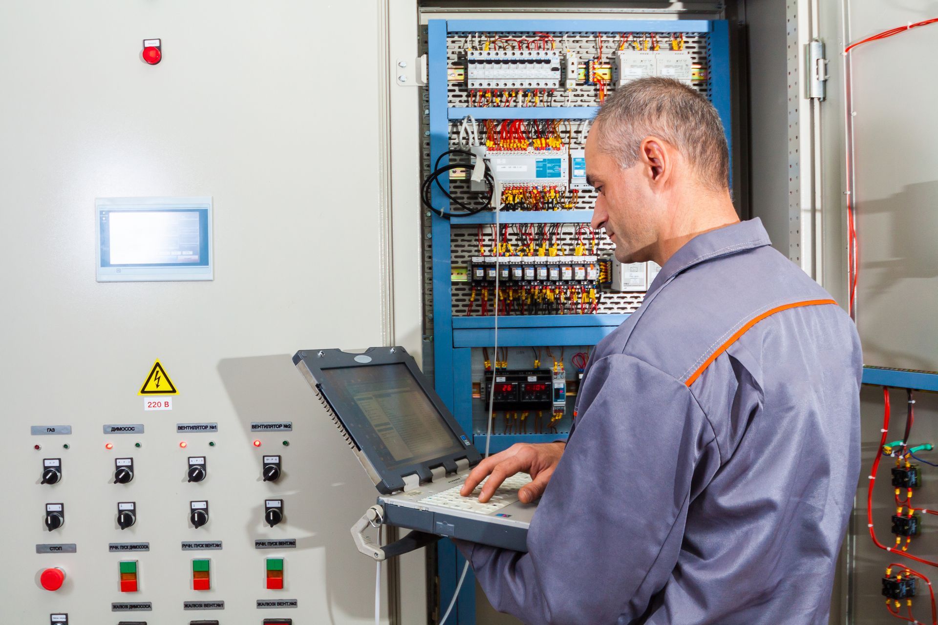 Man in work clothes uses a laptop to inspect an electrical control panel.