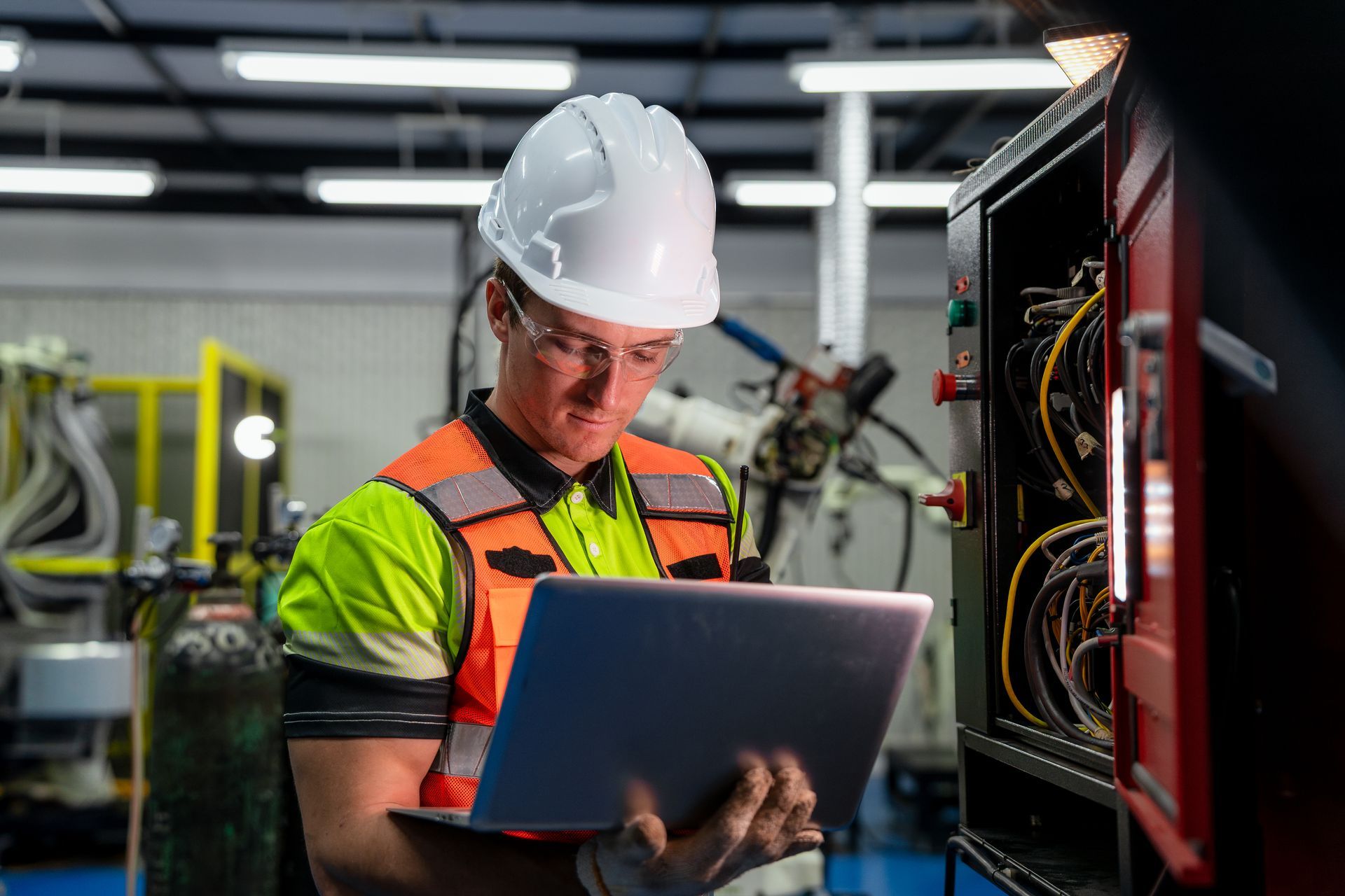 Engineer in a hard hat and safety vest using a laptop to inspect machinery in a factory.