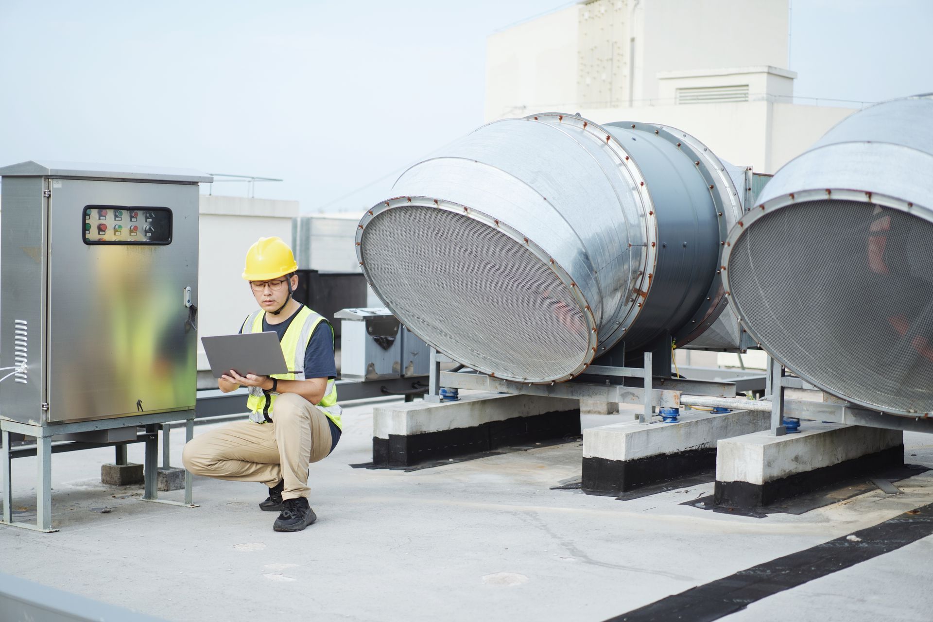 Engineer in hard hat inspects HVAC system on a rooftop, using a laptop near metal ventilation equipment.