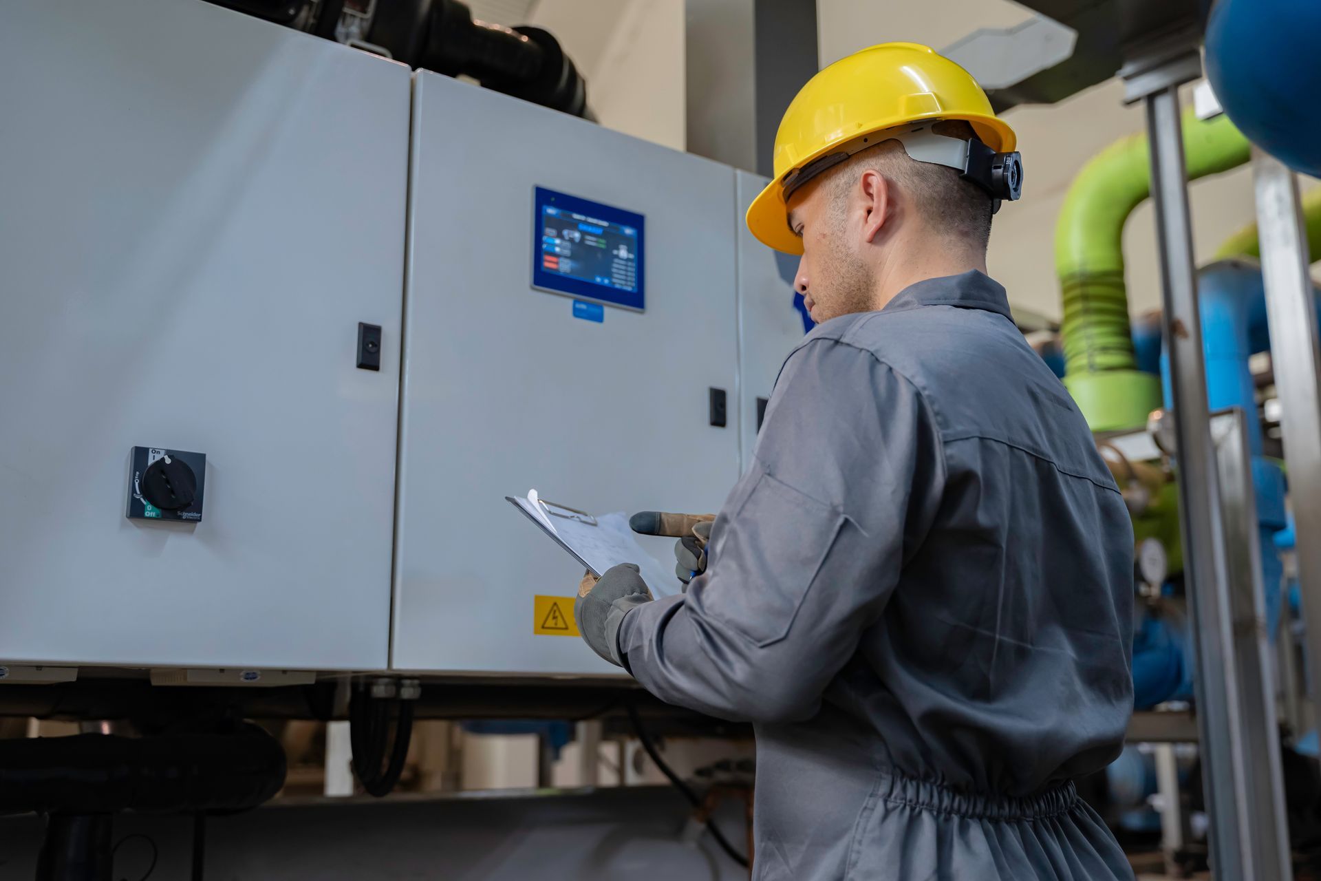 A person in a yellow hard hat and work clothes examines machinery, holding a clipboard in an industrial setting.