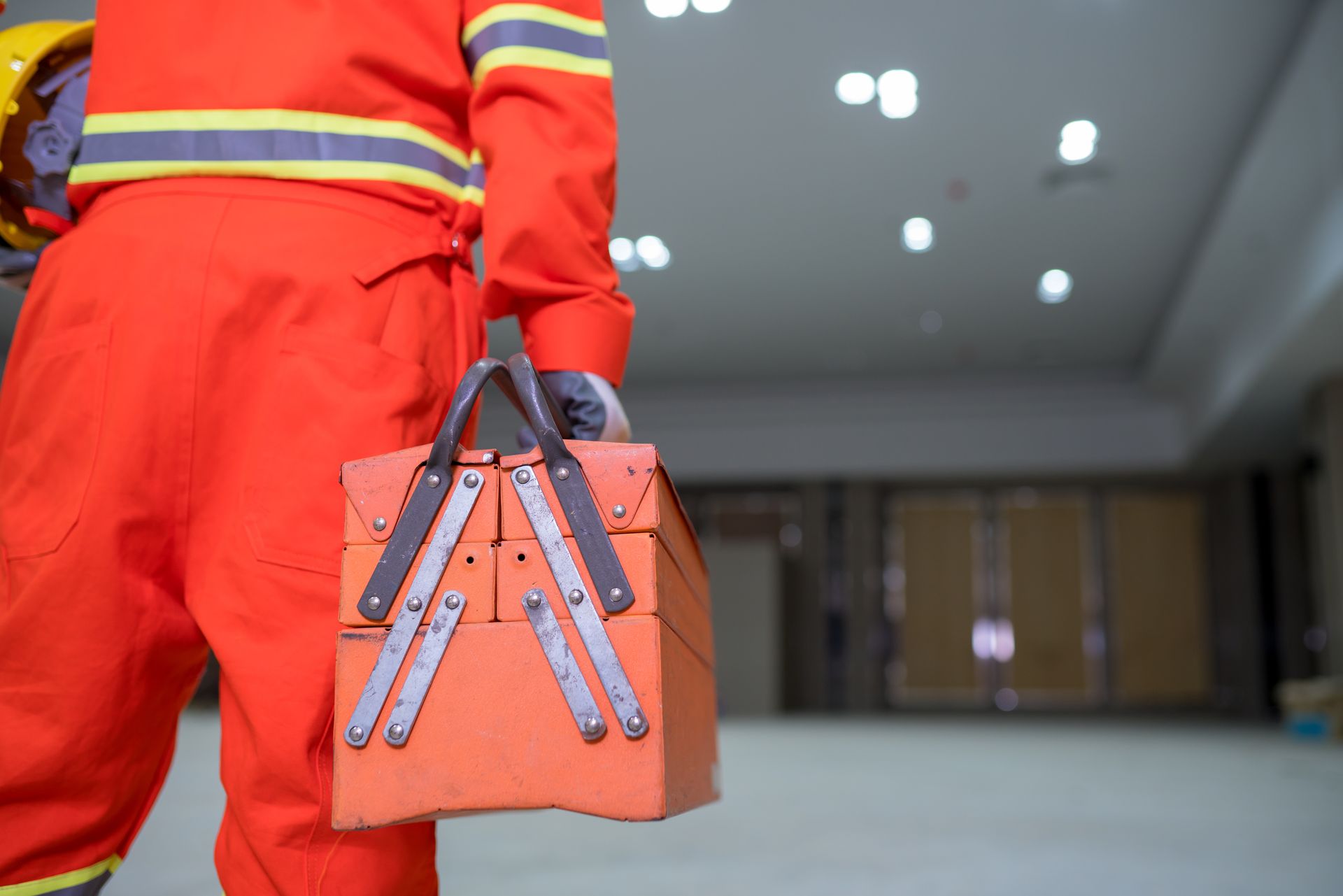 Worker in orange jumpsuit carrying an orange toolbox in a construction area.
