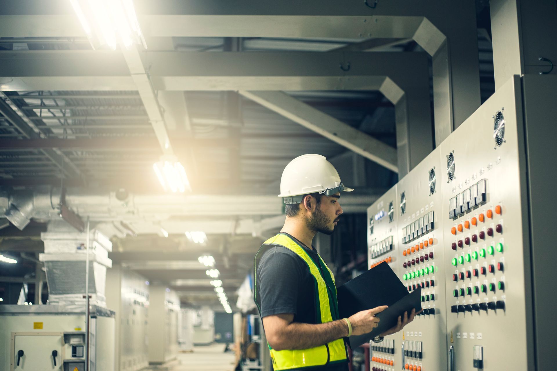Technician in hard hat and safety vest examines control panel in industrial setting.