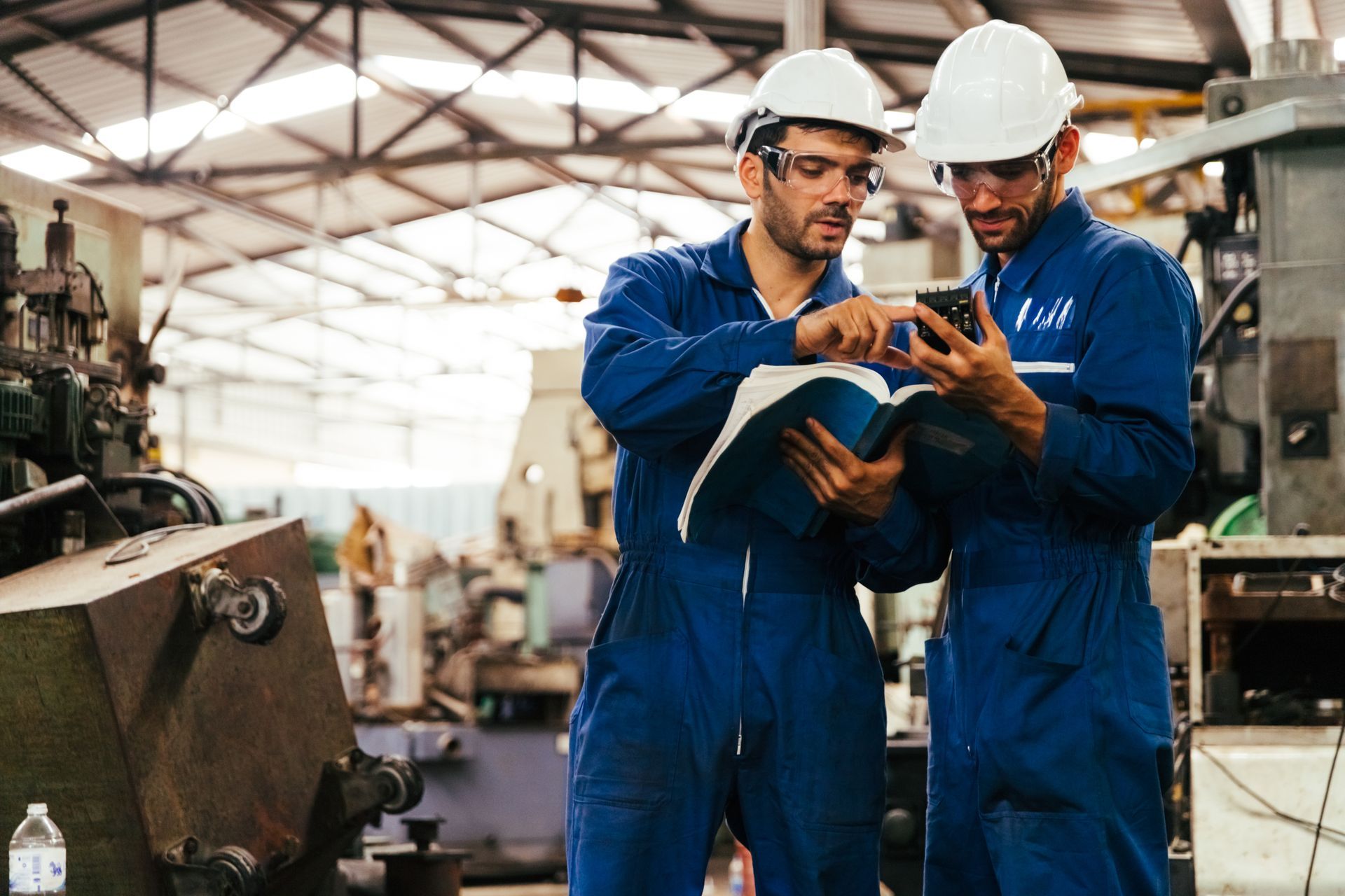 Two workers in blue jumpsuits and hard hats reviewing a document and phone in a factory setting.