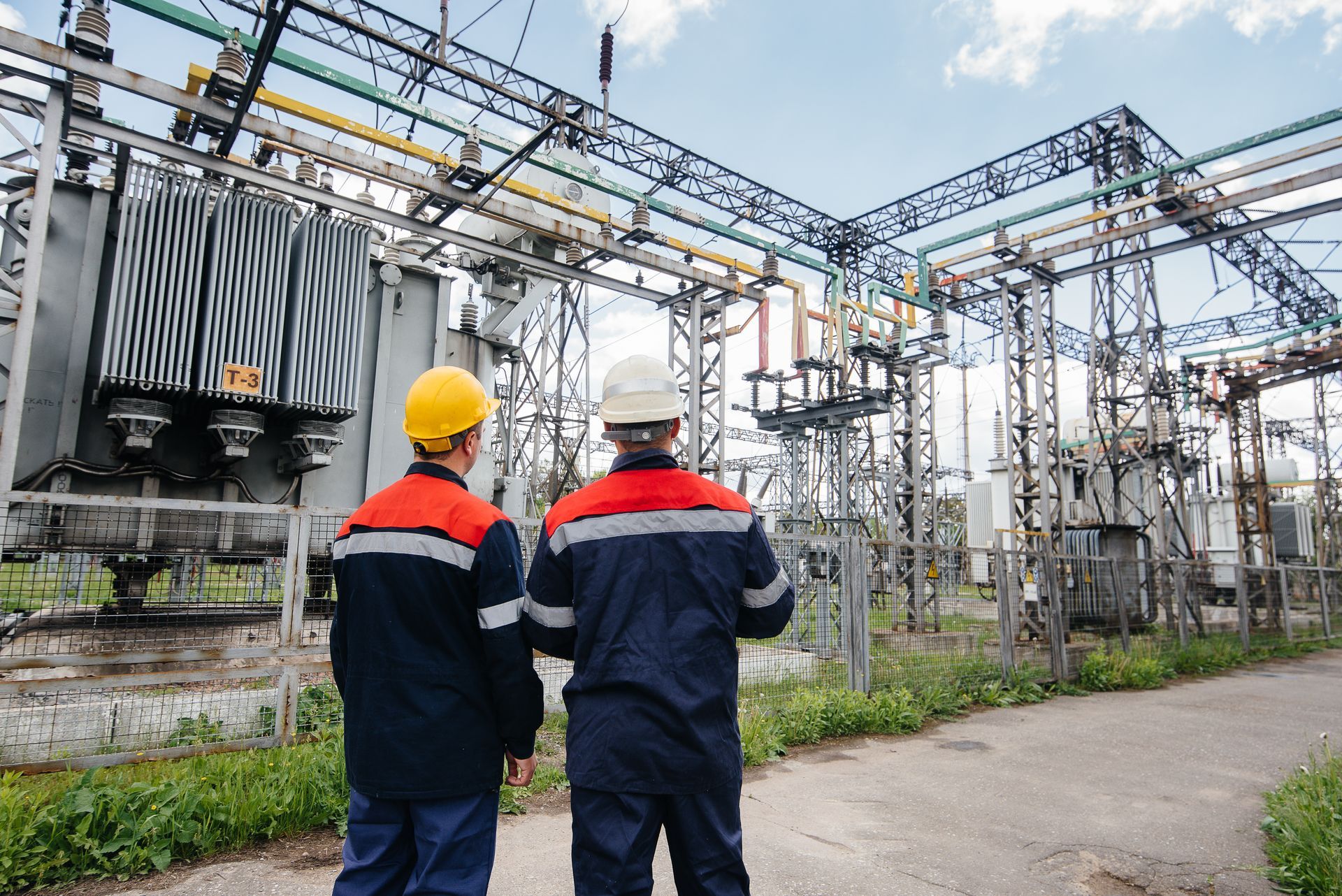 Two workers in safety gear at an electrical substation, inspecting equipment.