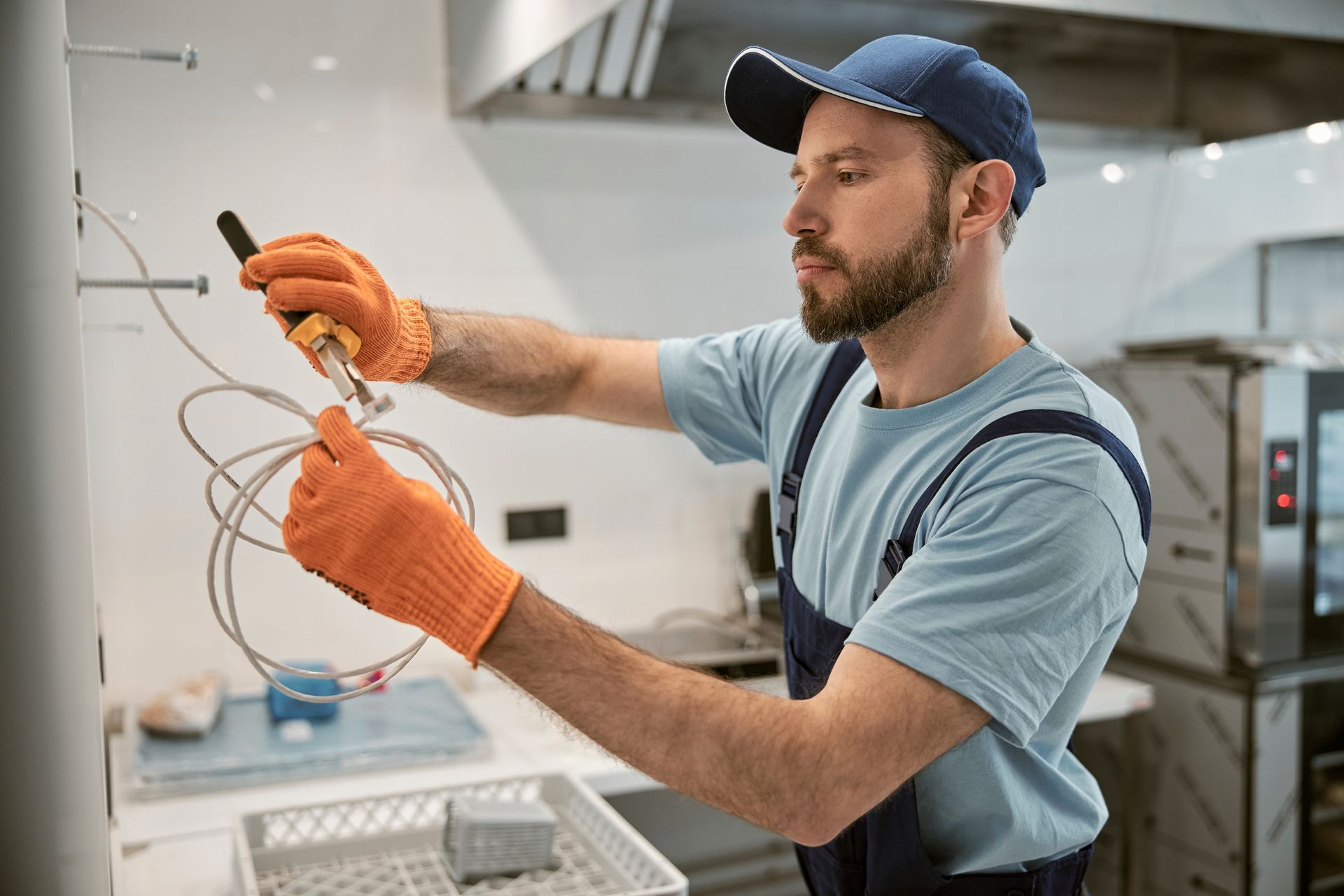 Man in overalls and cap using pliers to work with wires in a kitchen.