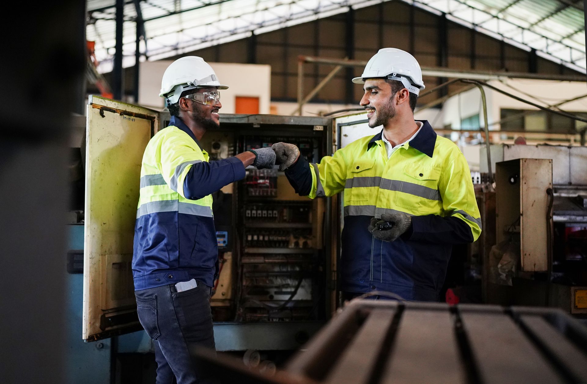 Two workers in safety gear fist bump near industrial equipment.