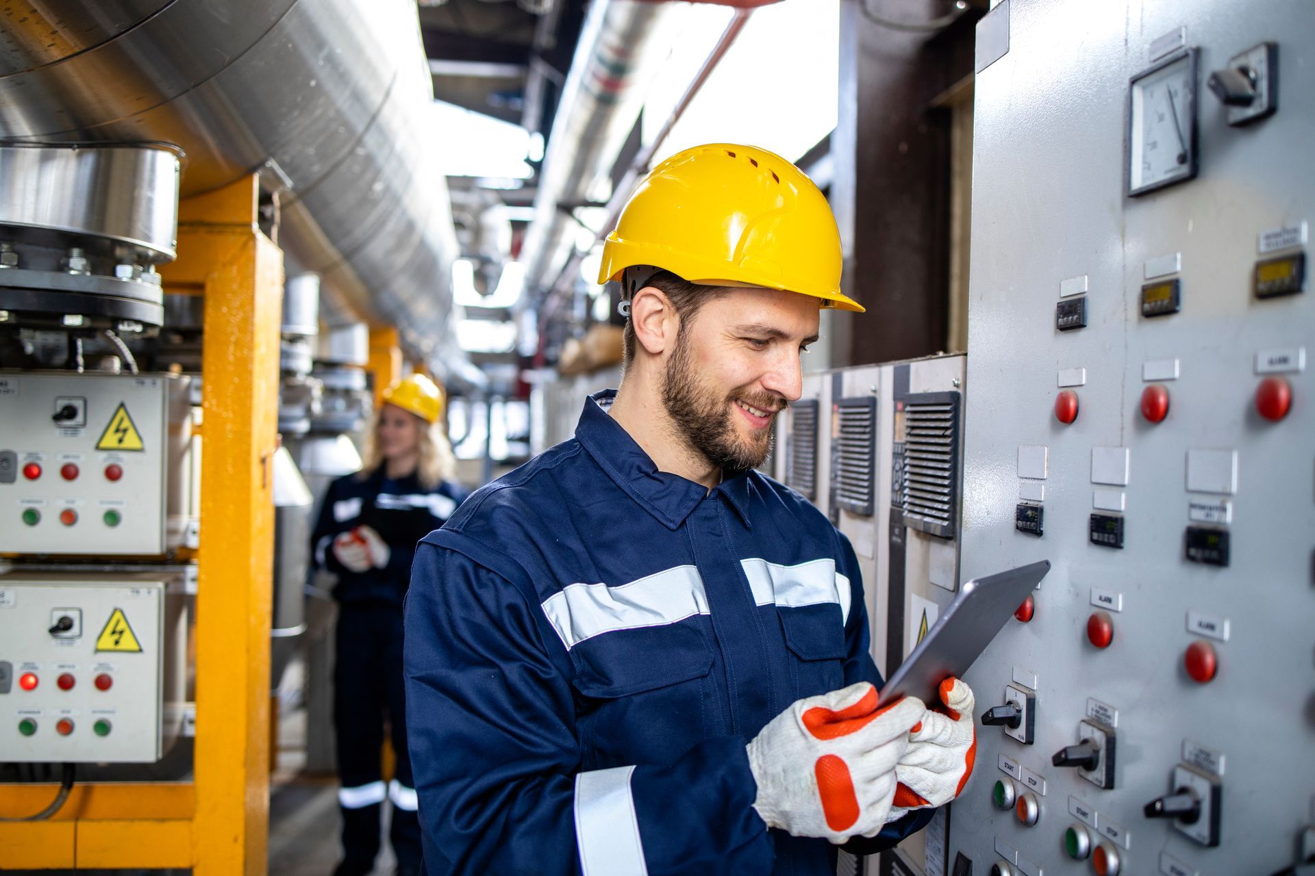Man in blue work suit and hard hat using a tablet near machinery; another worker in the background.