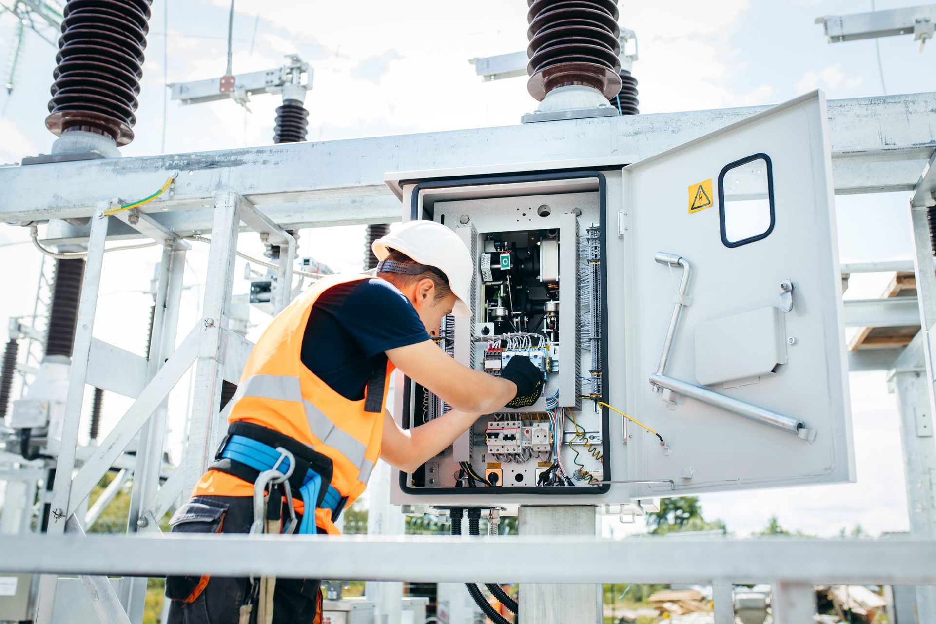 Electrician in a safety vest and hardhat working on electrical panel outdoors.
